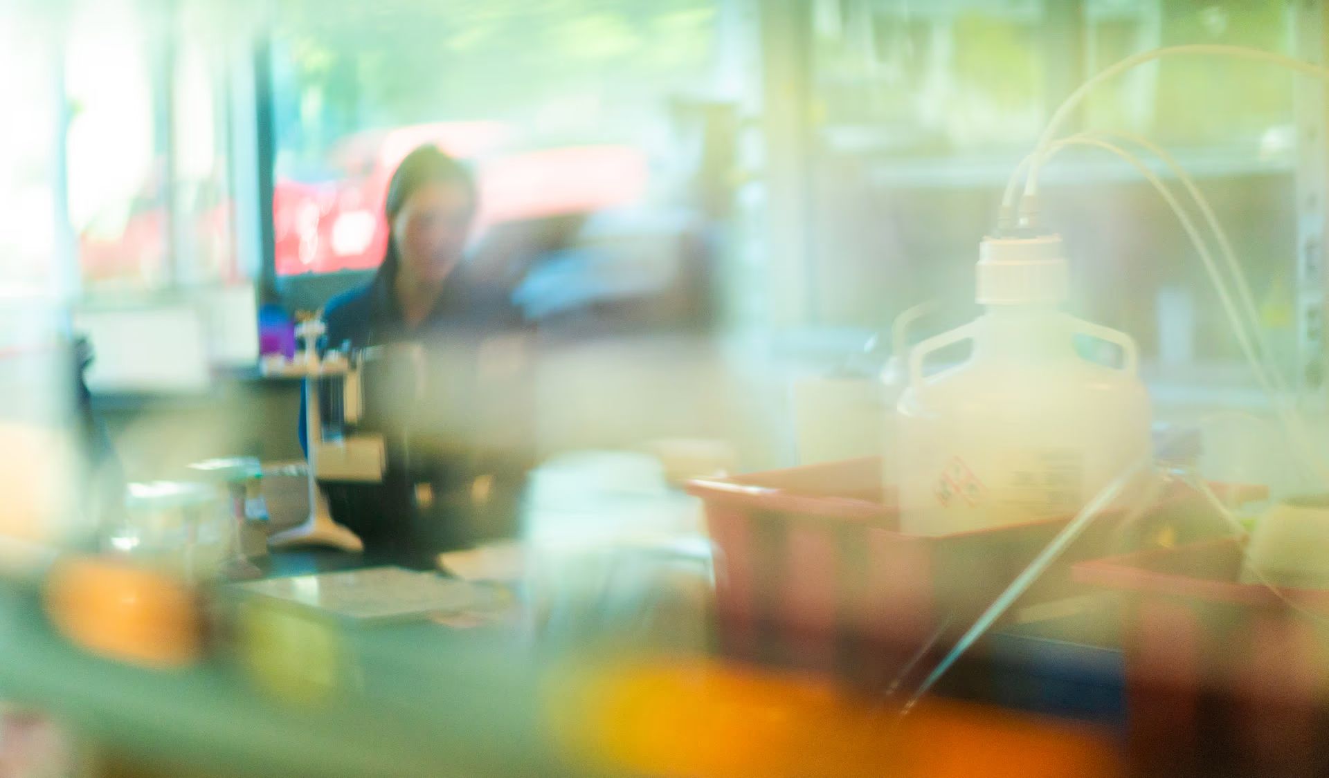A laboratory bench seen through glass, a researcher out of focus at a monitor behind carboys and trays, the whole frame washed in green and amber light