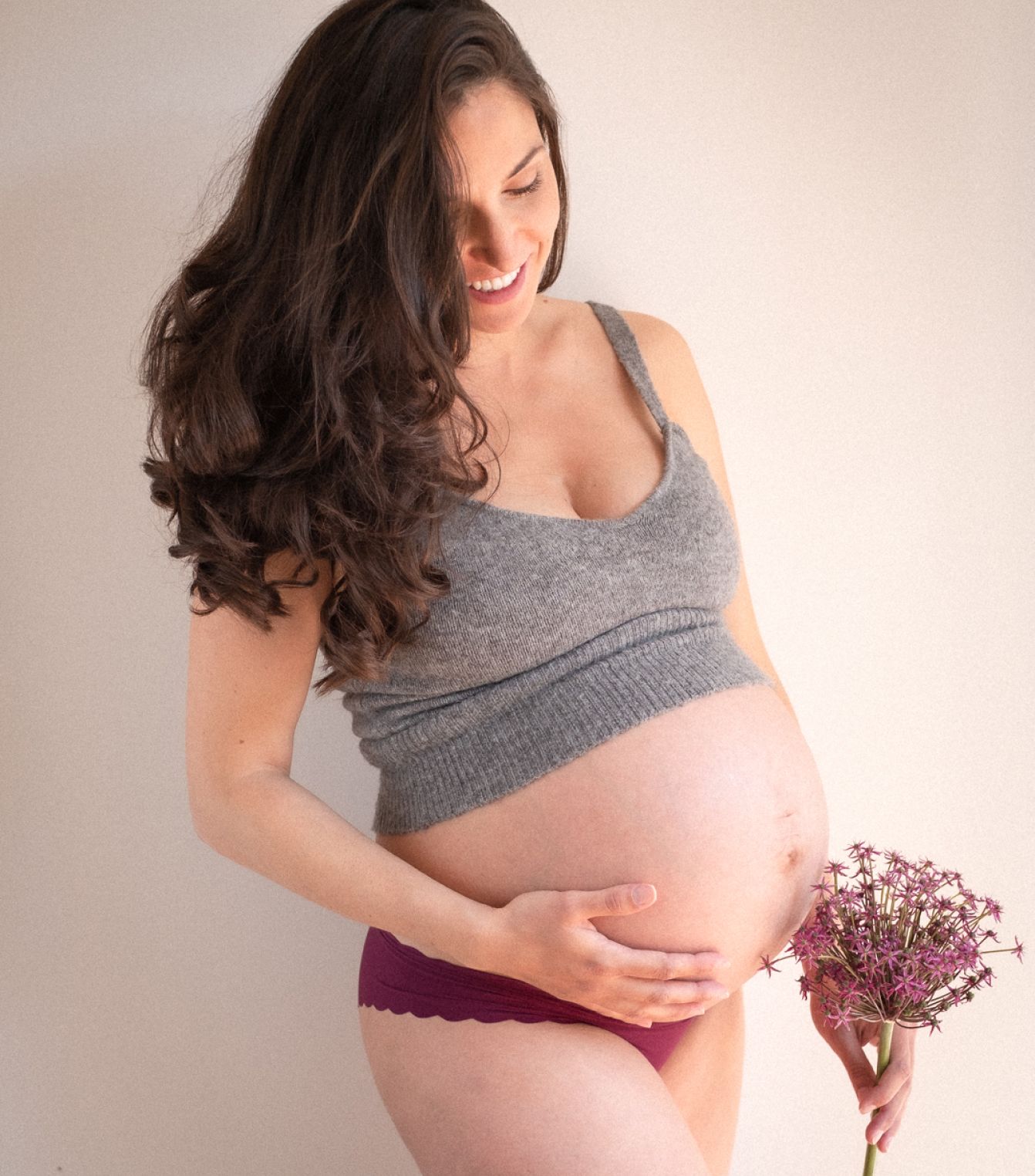 A pregnant woman in a grey knit top cradling her bump, an allium flower head in her other hand